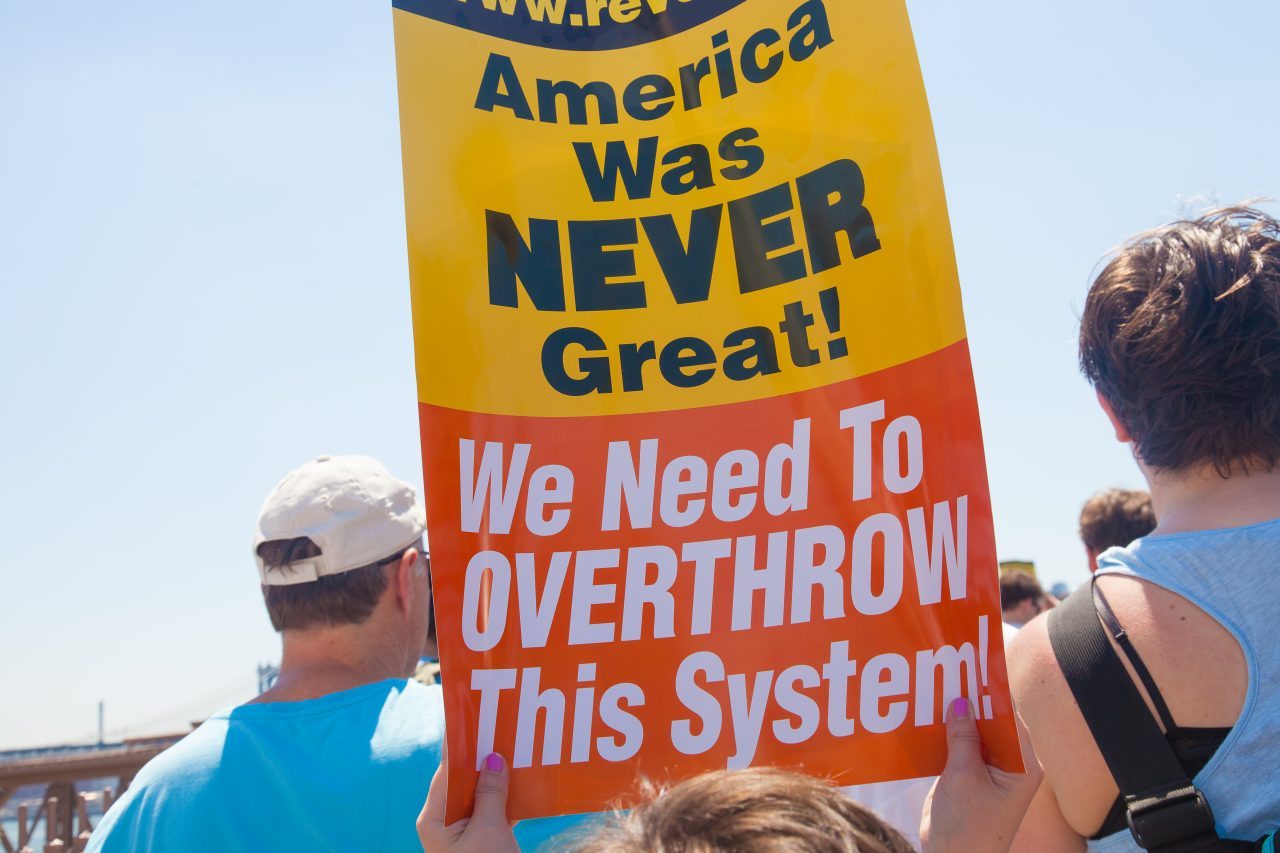 6/30/2018. New York City - March: “Families Belong Together.” A person with a sign that reads: "America was never great! We need to overthrow this system!" People crossing the Brooklyn Bridge in support of refugee families. People’s protectors are marching from Foley Square in Manhattan to Cadman Plaza in Brooklyn. Credit: Photo by Javier Soriano/LoveIsAmor.com